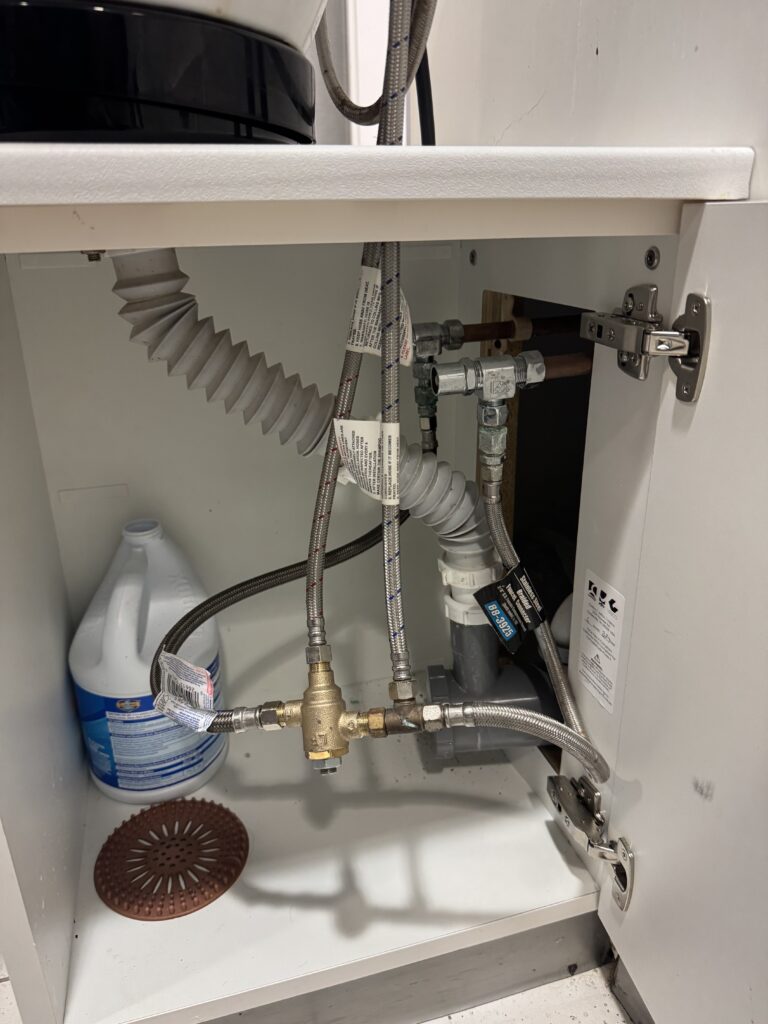 Plumbing setup under a sink with pipes, hoses, and cleaning supplies.