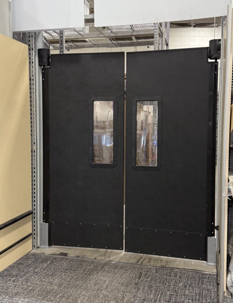 Black double doors with small glass windows in an industrial setting.